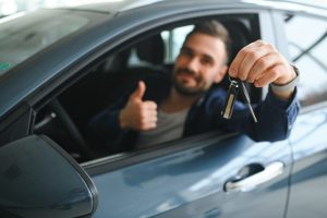 man inside car holding keys in one hand out the window and giving thumbs up with the other hand