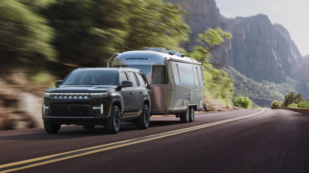 Black Jeep SUV towing a shiny silver Airstream trailer on a winding mountain road surrounded by trees and cliffs.