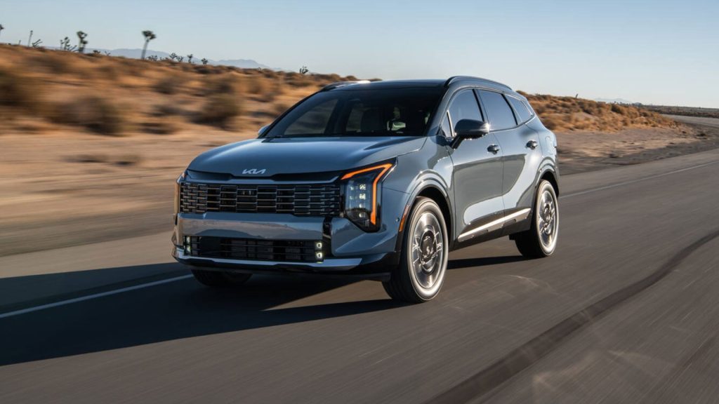 Silver Kia SUV driving on a desert highway with blurred brown shrubs and clear blue sky in the background.